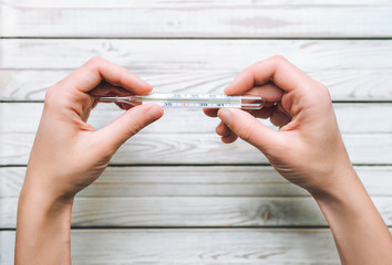 Increased temperature. Woman hands with mercury thermometer on white wooden background. The concept of cold, flu, acute respiratory diseases. Close up.