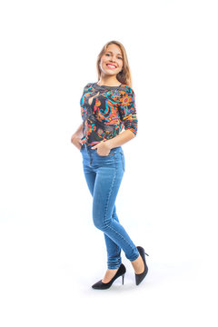 Attractive young woman with long blond hair in jeans, a blouse and shoes posing at the camera in profile with her hands in the pockets of jeans smiling.