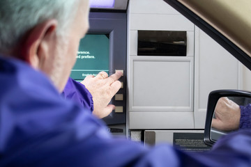 Grey-haired man at drive-up ATM machine leaning out car window and pushing button - Instructions in English and Spanish - Selective focus.