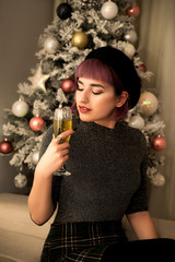 Young woman sitting near Christmas tree and drinking champagne