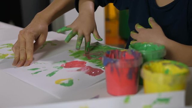 Close-up of asian preschool girl's hands stained in colorful paints doing hand painting on white paper with help of female educator at art class in kindergarten. Kid drawing with finger at art lesson.