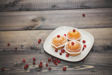 Freshly baked muffin decorated with red berries. Dusted with icing sugar on a white plate. Light wooden tabletop with scattered red berries.
