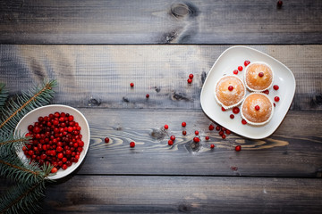 Fototapeta na wymiar A light wooden table top with a plate of freshly baked muffins decorated with red berries sprinkled with white powder and a plate of red berries with spruce twigs.