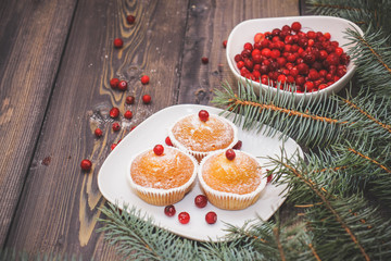 A light wooden table top with a plate of freshly baked muffins decorated with red berries sprinkled with white powder and a plate of red berries with spruce twigs.