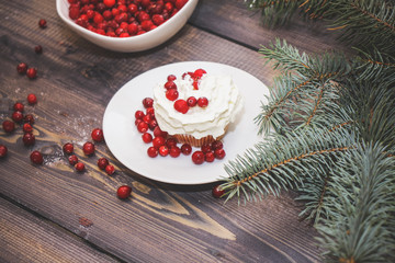 Freshly baked cake on a white plate decorated with red berries and whipped white cream with a plate topped with red berries decorated with spruce sprigs on a light wooden table top.