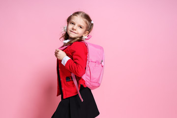 Back to school. Child with schoolbag. Schoolgirl 7-8 years old in a red jacket, white shirt, pigtails, black sarfan, school uniform with school bag isolated on pink
