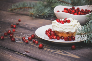 Freshly baked cake on a white plate decorated with red berries and whipped white cream with a plate topped with red berries decorated with spruce sprigs on a light wooden table top.
