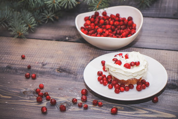 Freshly baked cake on a white plate decorated with red berries and whipped white cream with a plate topped with red berries decorated with spruce sprigs on a light wooden table top.