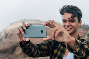 Young handsome guy making landscape photo on mobile phone, while standing on the mountain hill.