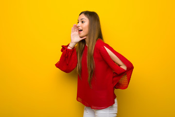 Young girl with red dress over yellow wall shouting with mouth wide open to the lateral