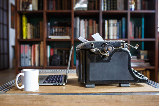 old vintage dust-covered typewriter with sheet of white paper near modern notebook and cup of coffee on bookcase background. modern technology and vintage appliances