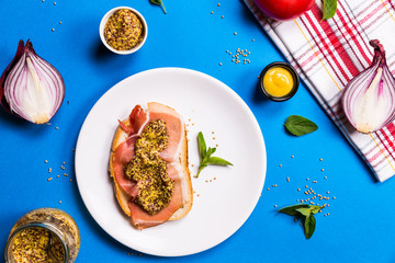 Top view of sandwich with ham and mustard on a white ceramic plate, blue backgrounds with ingredients