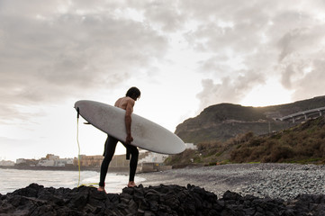 Side view brunette boy walking in the black swimsuit with naked torso with a white surf in his hands on the shore