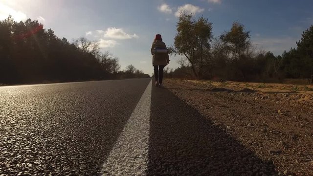 Young girl, tourist hitchhiker walks along road with a backpack on his shoulders, lifting his thumb up. Traveler is trying to stop car. Highway in summer forest