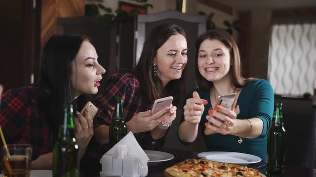 Girls pictures of pizza on your smartphone. The company of cheerful girls in the restaurant. Girls having fun drinking beer and eating pizza.