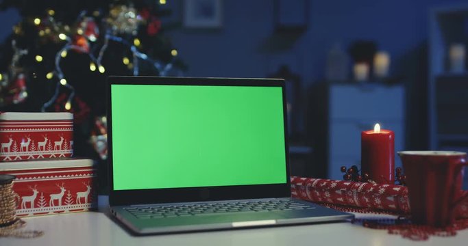 Close up of the laptop computer with green screen being on the table among wrapped Christmas presents in the decorated for x-mas dark home at night. Alpha channel. Chroma key. Indoor.