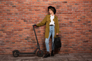 Young beautiful woman in trench coat,jeans and black hat holding backpack in hand while happily looking on electric scooter over brick wall background