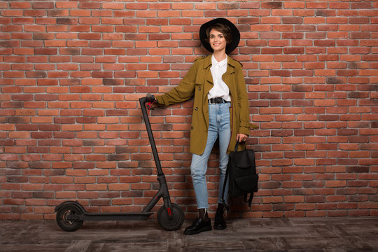 Young attractive woman in trench coat,jeans and black hat holding backpack in hand while happily looking in camera with electric scooter near over brick wall background