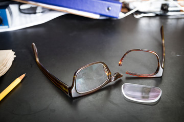 Broken eyeglasses on desk; lens of glasses fell out of broken frames.
