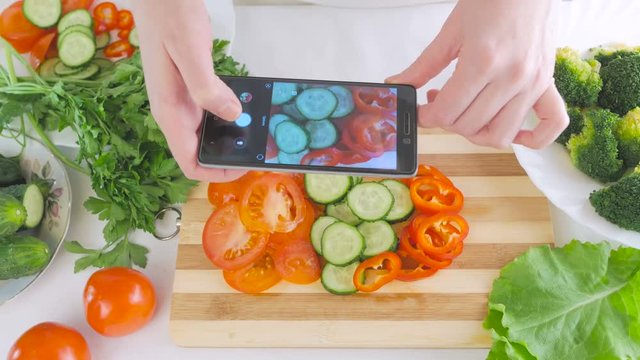 Woman's hands with a smartphone are taking pictures of fresh vegetables for salad, sliced on a cutting board.