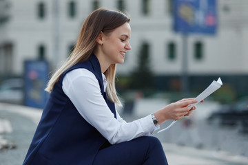 Pretty cheerful young blonde girl in smart casual eclectic outfit sits on cafe terrace while drinking coffee from paper cup. Coffee to go