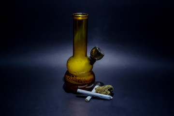 Fototapeta na wymiar An orange glass water bong full of smoke and a burning bowl with a small pile of Cheese OG marijuana buds next to it and 2 joints.