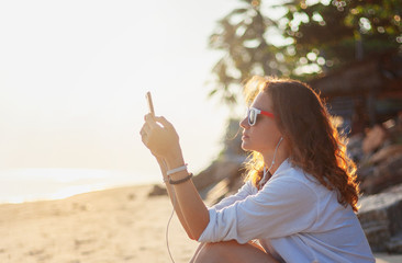 Beautiful young girl woman sitting on the beach at sunset in headphones and listening to music, rest and holidays