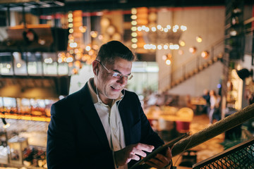 Close up of middle-aged smiling businessman dressed smart casual using tablet for video call while standing in cafeteria. In ears earphones.