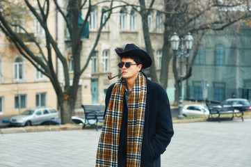 a man smokes a pipe in the city . portrait of a stylish fashionable man on the streets . man in black coat, plaid scarf and hat
