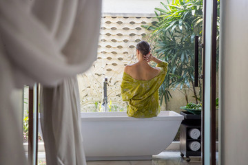 Back view of woman in a green bathrobe sitting at the tub edge in the bathroom, spa weekend,...