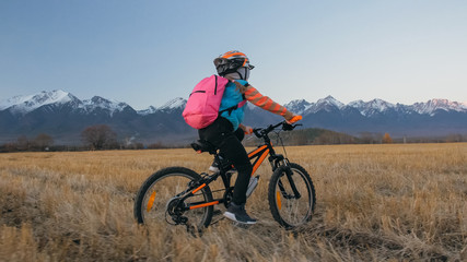 Fototapeta premium One caucasian children rides bike in wheat field. Little girl riding black orange cycle on background of beautiful snowy mountains. Biker motion ride with backpack and helmet. Mountain bike hardtail.