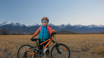 Fototapeta premium One caucasian children walk with bike in wheat field. Little girl walking black orange cycle on background of beautiful snowy mountains. Biker stand with backpack and helmet. Mountain bike hardtail.