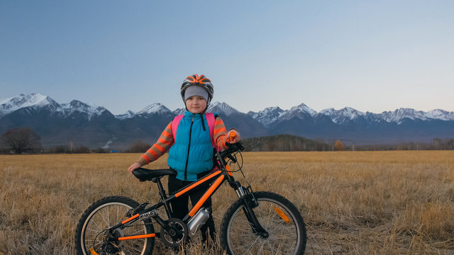 One caucasian children walk with bike in wheat field. Little girl walking black orange cycle on background of beautiful snowy mountains. Biker stand with backpack and helmet. Mountain bike hardtail.