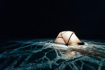 Fire on ice at night. Campground on ice. Tent stands next to bonfire. Lake Baikal. Nearby there is car. Shelter tent and ice are illuminated from the inside. Beautiful bonfire on real cracked ice