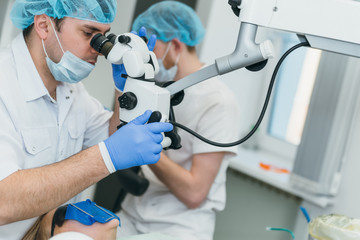 Doctor used microscope. Dentist is treating patient in modern dental office. Operation is carried out using cofferdam. Client is inserted and restored teeth, make denture. Orthodontist and assistant