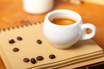 A Cup of espresso with foam and a notebook, scattered coffee beans on a wooden table. Coffee break concept