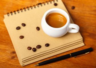 A Cup of espresso with foam and a notebook, scattered coffee beans on a wooden table. Coffee break concept