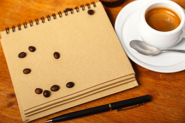 White Cup of fragrant espresso coffee with foam and writing pad, scattered coffee beans on a wooden table, copy space.