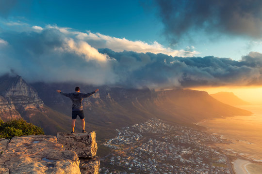 Young man standing on the edge at the top of Lion's head mountain in Cape Town with a beautiful sunset view