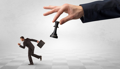 Fototapeta premium Small frail businessman with suitcase running away from big hand with chessman concept 