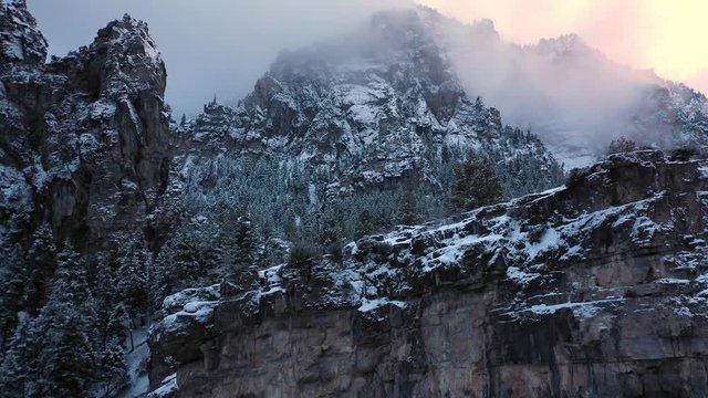 Panning aerial view of cliff in winter forest landscape as the sun is setting and clouds cover the mountain tops.