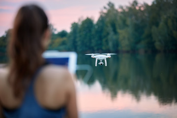 Young beautiful woman controls a white quadcopter by remote controller with a tablet. Focused on the drone, the pilot is out of focus. Teaching flying with a dron in summer nature.