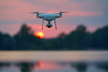 Quadcopter model hovering over lake against blurred red setting sun in background.