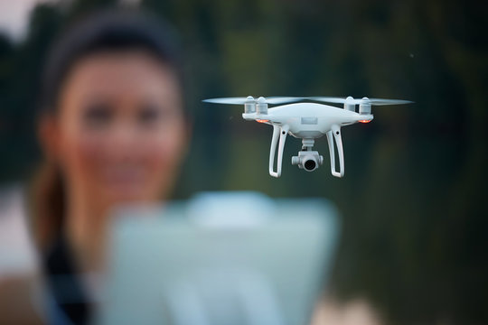 Young beautiful woman controls a white quadcopter by remote controller with a tablet. Focused on the drone, the pilot is out of focus. Teaching flying with a dron in summer nature.