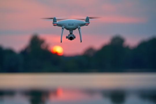 Quadcopter model hovering over lake against blurred red setting sun in background. - Powered by Adobe