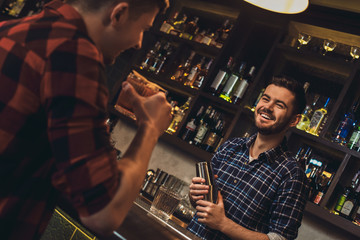 Young bartender standing at bar counter holding shaker talking with customer cheerful