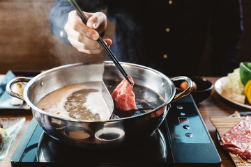 Man holding rare slice Wagyu A5 beef into shabu hot pot shoyu soup base by chopsticks with steam.