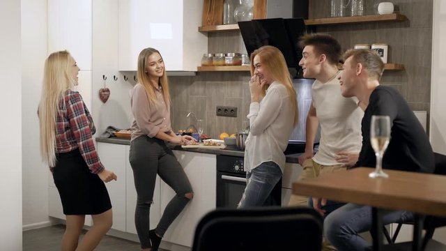 joyful female and male students are chatting on a kitchen during home party, smiling and laughing
