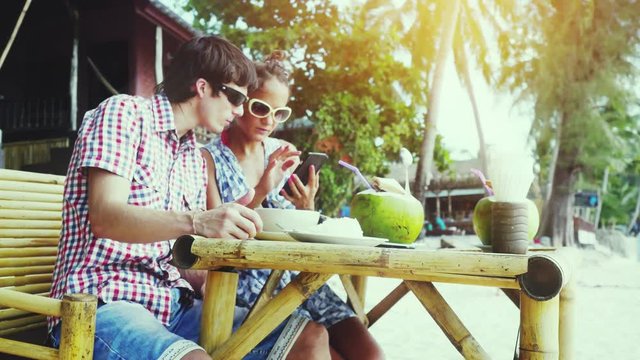 Pretty young couple in sunglasses sitting at a nice cafe by the beach. Girl showing man something in mobile phone. 3840x2160