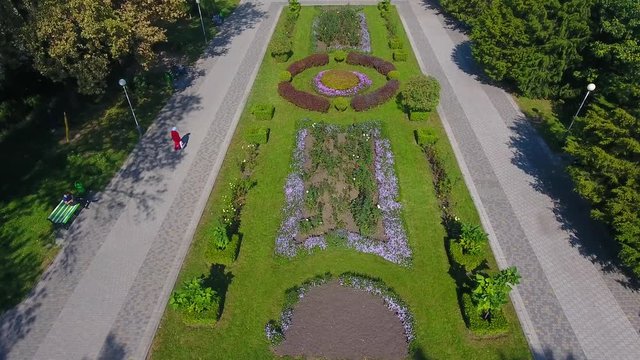 Aerial view: flower beds on hill in shade, people relax on the benches in the Park and people walk of many trees among in Maxim Gorky park to Krasnodar, cinematic aerial motion.